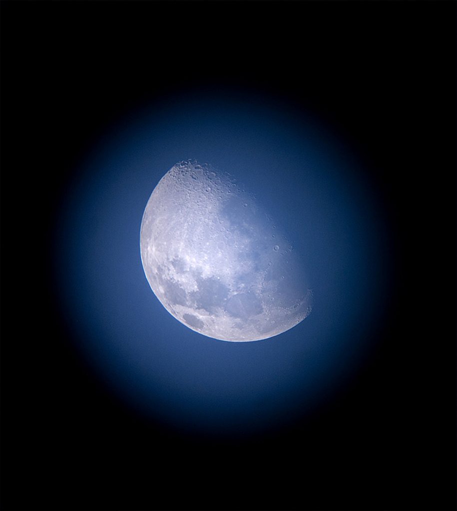 A daytime shot of the moon through a telescope, the background is clearly blue and there's a black halo around the Moon. It's very pale.